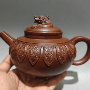 May include: A brown clay teapot featuring a rounded body with embossed petal-like patterns around the center. The lid is topped with a sculpted mythical creature figure. The teapot includes a curved spout, a circular handle, and three small feet.