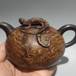 May include: A brown and yellow patterned teapot with a lizard-shaped lid handle. The teapot has a rounded body, a curved handle, and a spout. The lid features a detailed lizard sculpture. The teapot is held in a hand.