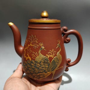 Puede incluir: Una tetera de cerámica marrón con tapa y asa doradas. La tetera está decorada con diseños dorados de pavos reales y flores. La boquilla y el asa de la tapa tienen detalles dorados.