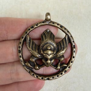 Può includere: Ciondolo color bronzo con un uccello stilizzato o creatura mitologica all'interno di una cornice circolare punteggiata. Il ciondolo ha un anello nella parte superiore per appenderlo. Il design è dettagliato e intricato.