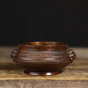 May include: A bronze-colored incense burner with a woven basket pattern. The small bowl has two lion head handles and sits on a round base. The background is a dark, solid color, and the burner is on a wooden surface.