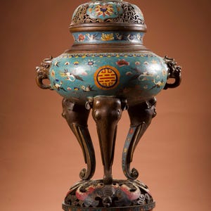 Op de afbeelding: Een sierlijke, antieke bronzen wierookbrander met een turquoise emaille afwerking. De brander heeft een gedetailleerd ontwerp met bloemen- en vogelmotieven, ondersteund door poten in de vorm van olifanten. Bovenop het deksel staat een klein bronzen hondenbeeld.