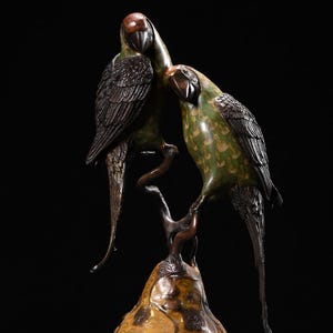 Peut inclure: Une sculpture en bronze de deux perroquets perchés sur une branche, avec une base en forme de poire. Les perroquets ont un plumage vert et marron, avec des détails de plumes. La base est décorée d'un motif floral doré et rouge.
