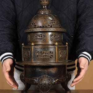 Op de afbeelding: Een bronzen wierookbrander met ingewikkeld opengewerkt en een gewelfd deksel. De brander heeft een gelaagd ontwerp met gedetailleerde reliëfsnijwerk en staat op drie sierlijke poten. De algehele kleur is een donker, verouderd brons.