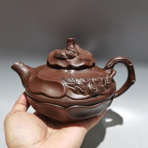 May include: A brown ceramic teapot with a floral design on the lid and body. The teapot has a curved handle and a spout. The teapot is held in a hand.