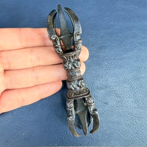 Pode incluir: Um objeto ritual vajra de metal cinza-escuro com um cabo central com entalhes em relevo intrincados e duas extremidades simétricas, cada uma composta por quatro pontas curvas que se encontram num ponto central, criando um design equilibrado.