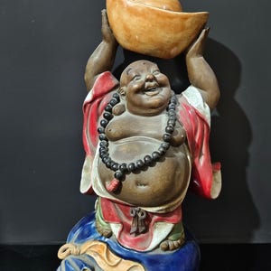 以下が含まれることがあります： 笑顔の仏陀が頭上に金塊を持っている陶器の像。仏陀は白いトリムの赤いローブと黒いビーズのネックレスを身に着けています。像は金のアクセントが付いた青い台座の上にあります。