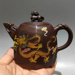 May include: A brown ceramic teapot featuring a golden dragon design with red and green accents. The teapot has a small animal figure on the lid and detailed spout and handle. The teapot is held in a person's hand.