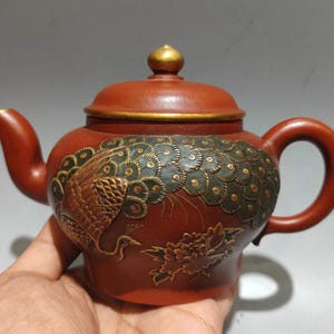 Puede incluir: Una tetera de cerámica marrón rojizo con un diseño de pavo real. La tetera presenta una tapa y detalles dorados, un pico curvo y un asa redondeada. El diseño del pavo real está detallado con detalles verdes y dorados, y también se ve un motivo floral.