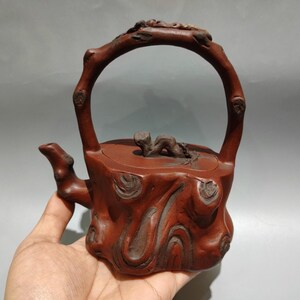 Op de afbeelding: Een roodbruine theepot met een grote handgreep en een tuit. De theepot is gesneden om op hout te lijken, met zichtbare knopen en nerfpatronen. De handgreep is gebogen en het deksel heeft een klein decoratief element.