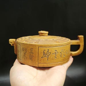 Könnte beinhalten: Hellbraune, handgefertigte Teekanne mit Deckel und Henkel. Die Teekanne hat eingravierte chinesische Schriftzeichen und ein geometrisches Muster auf dem Deckel. Die Teekanne wird in einer Hand vor dunklem Hintergrund gehalten.