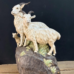 Op de afbeelding: Een keramieken sculptuur met twee volwassen geiten en een klein geitje op een gestructureerde, donkerbruine rotsformatie met groene grasaccenten. De geiten zijn gebroken wit met donkerbruine hoorns en gedetailleerde vachtstructuren.