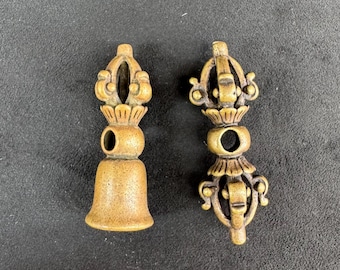 May include: Two brass-colored metal objects with intricate, symmetrical designs. One object features a bell-shaped base topped with a multi-pronged finial, while the other is a double-ended vajra with a central circular opening and ornate prongs.