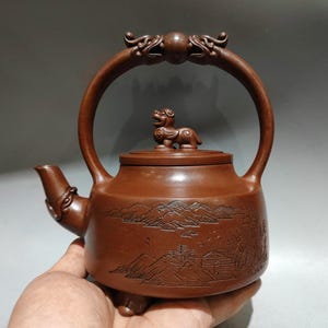 Puede incluir: Tetera de cerámica marrón con un asa curva y una pequeña figura de animal en la tapa. La tetera presenta detalles de paisaje tallados y un pico decorativo. El asa está adornada con tallas.