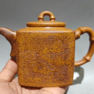 Puede incluir: Tetera pequeña, de forma cuadrada, con tapa y asa de bambú. La tetera es de color marrón cálido y terroso, con una superficie texturizada y un diseño de paisaje. El pico es curvo y el asa arqueada.