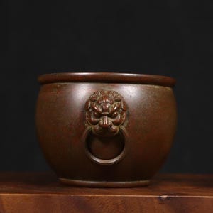 May include: A bronze-colored, round bowl with a lion's head and ring handle. The bowl sits on a wooden surface against a dark background. The lion's head is intricately detailed, adding a touch of artistry to the piece.