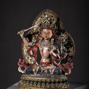 Könnte beinhalten: Eine mehrfarbige Statue einer Gottheit, die in einer Lotussitzposition auf einem Sockel sitzt. Die Figur trägt eine verzierte Krone, Schmuck und rote Bänder, hält ein Schwert in der rechten Hand und eine Lotusblume mit einem Buch in der linken Hand, vor einem großen, kunstvollen, goldfarbenen Heiligenschein.