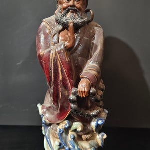 May include: A detailed ceramic statue of a wise-looking man with a long beard and a serene expression. The figure wears a brown robe with red accents and holds prayer beads. The base features a wave design.