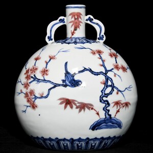 Puede incluir: Un jarrón de porcelana azul y rojo con dos asas. El jarrón está decorado con un pájaro posado en una rama florida. El jarrón tiene un cuerpo redondo y un cuello estrecho.