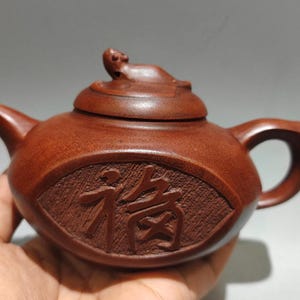 May include: A small, brown wooden teapot with a curved handle and spout. The lid features a small, decorative element. The body of the teapot has carved Chinese characters. The teapot is held in a hand.