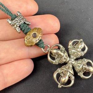 May include: A silver pendant with a four-lobed design, attached to a green cord. The pendant features detailed patterns and is likely metal. The cord has a decorative clasp and a brass-coloured bead.