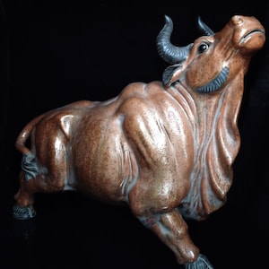 Può includere: Scultura di toro in ceramica marrone con finitura lucida. Il toro è raffigurato con la testa alzata, mostrando corna nere ricurve. La scultura presenta una muscolatura dettagliata ed è ambientata su uno sfondo nero.