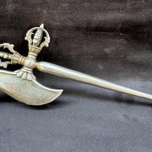Puede incluir: Un cuchillo ritual plateado con una hoja curva y un mango ornamentado. El mango presenta diseños intrincados y un elemento en forma de corona. La hoja tiene forma de media luna, lo que sugiere un uso ceremonial o decorativo.