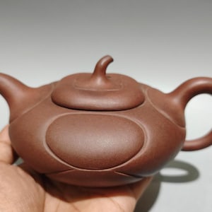 Puede incluir: Una tetera de arcilla marrón con acabado mate, que presenta un cuerpo redondeado con paneles ovalados en relieve, un pico curvo, un asa circular y una tapa rematada con un pequeño remate curvo. La tetera tiene una textura suave y uniforme.