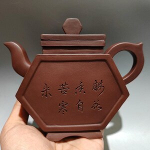 Op de afbeelding: Roodbruine keramische theepot met een zeshoekige body en een rechthoekig deksel. De theepot heeft een gebogen tuit en een handvat met een cirkelvormige opening. Chinese karakters zijn op de voorkant gegraveerd.