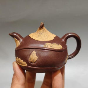 May include: A dark brown clay teapot featuring a textured, light tan lid with a pointed finial. The body of the teapot is decorated with raised, light tan, cloud-like shapes and teardrop patterns, complemented by a curved handle and a short spout.