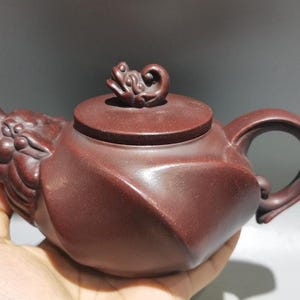 May include: A maroon-colored teapot with a dragon head spout and a frog-shaped handle on the lid. The teapot has a curved handle and a geometric design on the body. The teapot is held in a hand.