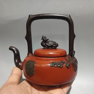 Puede incluir: Una tetera de color rojo anaranjado con un asa y un pico de color marrón oscuro. La tapa presenta una pequeña figura de dragón y tortuga de color oscuro. Acentos florales decorativos de color bronce adornan el lateral de la tetera. El asa tiene forma arqueada.