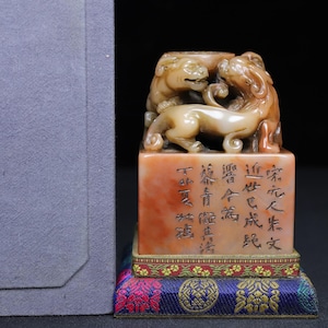 May include: A rectangular stone seal featuring a carved mythical creature on top. The base is orange and beige with engraved Chinese characters. It rests on a decorative fabric stand with blue, red, and gold patterns, positioned beside a grey box.