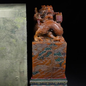 May include: A carved amber-coloured stone seal featuring a dragon sculpture on top and a landscape relief of a pine tree on the side. The seal rests on a multi-layered base with geometric patterns and is accompanied by a tall, light-coloured storage box.