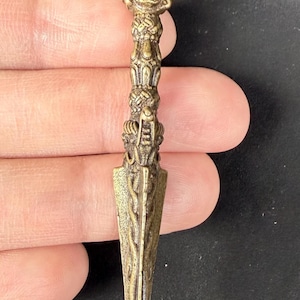 May include: A detailed, antique-style dagger with a bronze finish. The handle features intricate carvings, including a detailed face at the top. The blade is pointed and has a textured surface, showcasing a traditional design.