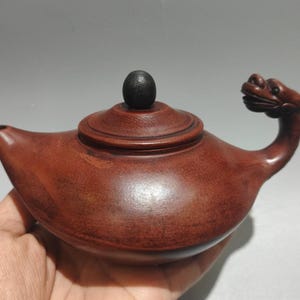 May include: A reddish-brown wooden teapot with a dragon-shaped handle and a round lid. The teapot has a small spout and a black knob on the lid. The teapot is held in a hand.