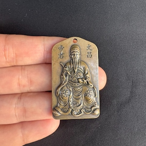 Könnte beinhalten: Bronzefarbener rechteckiger Anhänger mit einer sitzenden Figur mit langem Bart und traditioneller Kleidung. Der Anhänger hat chinesische Schriftzeichen oben und ein Loch zum Aufhängen. Die Figur ist aufwendig detailliert.