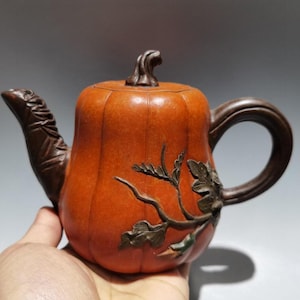 Puede incluir: Una tetera con forma de calabaza naranja con asa, boquilla y tapa marrones. La tetera presenta un diseño floral tallado. La tetera se sostiene en una mano.