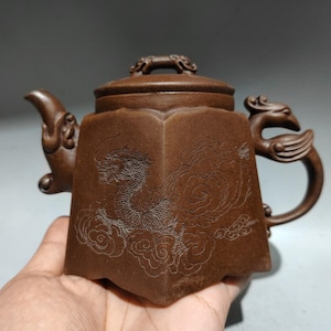 Op de afbeelding: Een bruine kleien theepot met een zeshoekig lichaam voorzien van een gegraveerd drakenontwerp tussen wolken. De theepot heeft een deksel met een kleine handgreep, een gebogen tuit en een handvat in de vorm van een gestileerde vogel.
