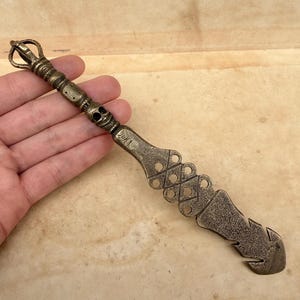 May include: A bronze-coloured metal butter knife with a decorative handle featuring a skull and ornate patterns. The knife has a unique, textured design with a curved end and cut-out details, suitable for a kitchen or dining setting.