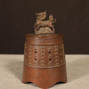 May include: A cylindrical bronze-toned metal incense burner featuring a textured surface with vertical rows of raised studs and small circular perforations. The lid is topped with a detailed sculptural figure of a mythical creature.