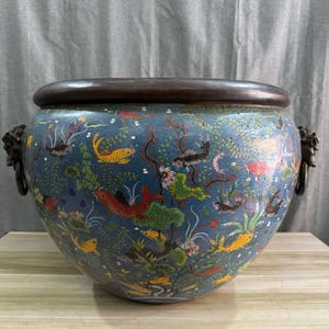 Peut inclure: Un grand pot en céramique bleu rond avec un bord en bronze et des poignées en tête de lion. Le pot est décoré de poissons colorés et de plantes aquatiques. Les poissons sont rouges, jaunes et noirs. Les plantes sont vertes et blanches.