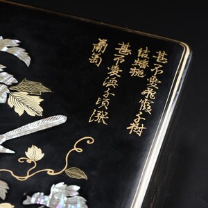 May include: A black rectangular box featuring an intricate inlay of iridescent mother-of-pearl depicting grapevines and a bird. The surface is decorated with gold-colored calligraphic text and delicate gold-colored vine outlines.