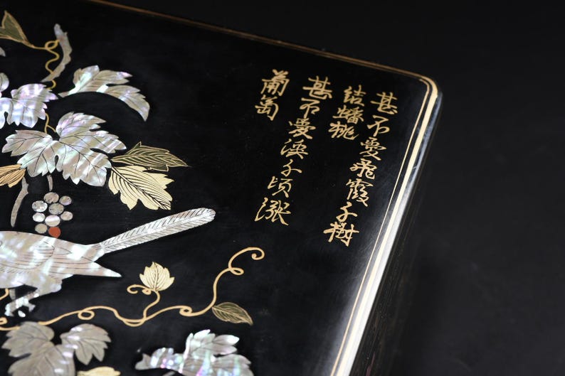 May include: A black rectangular box featuring an intricate inlay of iridescent mother-of-pearl depicting grapevines and a bird. The surface is decorated with gold-colored calligraphic text and delicate gold-colored vine outlines.