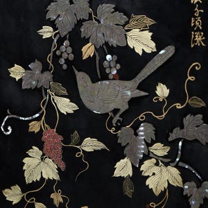 May include: A black panel featuring an inlaid design of a bird perched among grapevines. The artwork uses iridescent mother-of-pearl for the bird and leaves, with red accents for the grapes and gold-colored line work. Chinese characters appear in the top right corner.