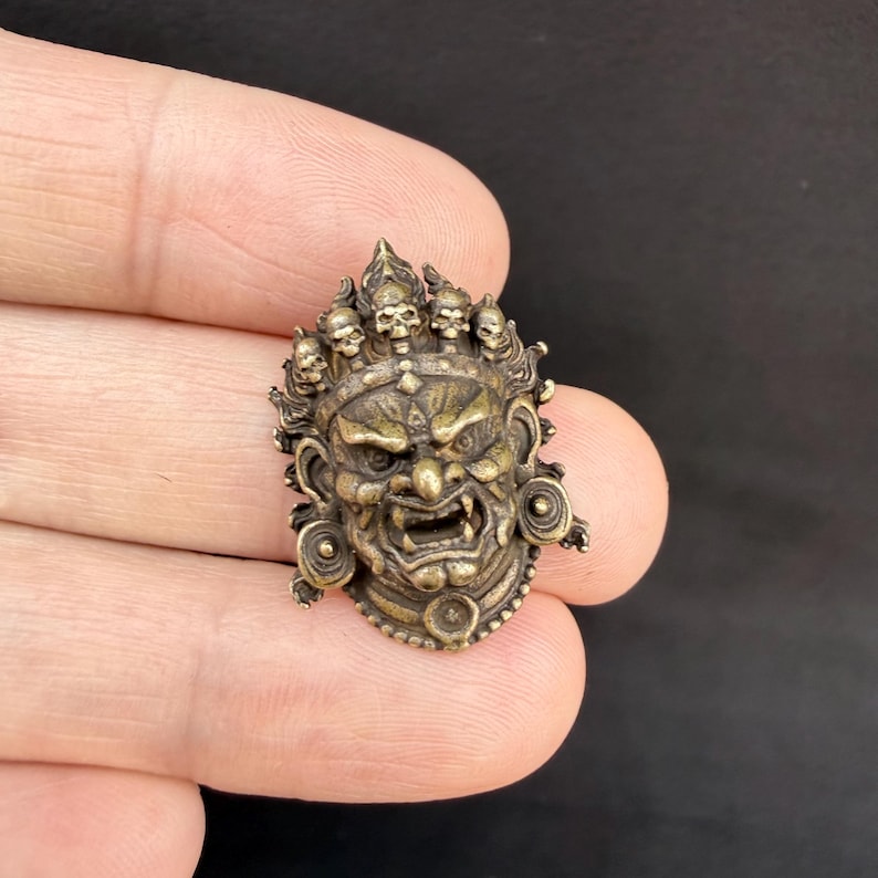 May include: A detailed bronze-colored pendant in the shape of a fierce face, adorned with a crown of small skulls. The face has an angry expression with bared teeth and intricate details. The pendant is held in a hand against a dark background.
