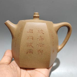 Könnte beinhalten: Eine hellbraune Keramik-Teekanne mit Henkel, Ausguss und Deckel. Die Teekanne hat eine achteckige Form und ist mit chinesischen Schriftzeichen beschriftet. Die Teekanne wird in der Hand einer Person gehalten.