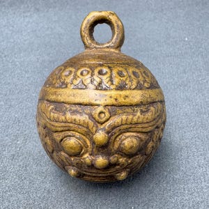 May include: A small, round, gold-colored bell with a detailed face and a loop at the top. The bell has a decorative design around the top edge.