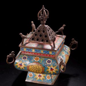 Puede incluir: Un quemador de incienso de metal antiguo y ornamentado, con una base cuadrada y una parte superior piramidal. El quemador está decorado con coloridos diseños florales en azul, amarillo y rojo. Pequeñas figuras de pájaros adornan la parte superior y asas de anillo en los lados.