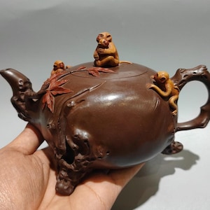 May include: A dark brown clay teapot featuring sculpted light brown monkeys on the lid and side. The teapot has a textured handle and spout resembling tree branches, with carved red maple leaves decorating the surface of the rounded body.
